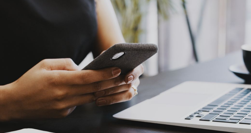 woman texting on cell phone next to laptop on desk
